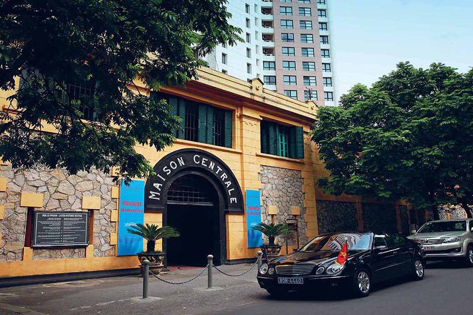 Ochre street facade of the Hoa Lo Prison building with cars parked outside
